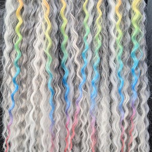 May include: A collection of wavy, gray hair extensions with vibrant rainbow-colored streaks. The streaks transition from orange to yellow, green, blue, and pink. The extensions are arranged in a row, showcasing the colorful highlights.