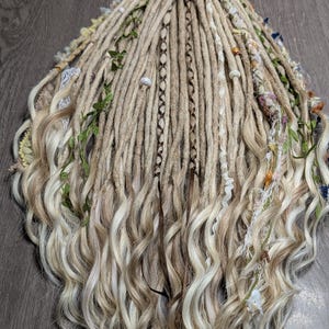 May include: Blonde and light brown dreadlocks with wavy ends, decorated with small flowers, green leaves, and decorative accents. The dreadlocks are arranged in a bundle, showcasing a natural, bohemian style.