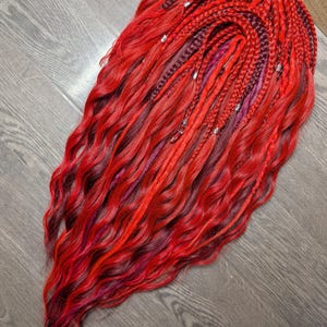 May include: A striking, wavy hairpiece in shades of red, burgundy, and purple. The hair is styled with braids and embellished with silver accents. The overall appearance is bold and striking, ideal for a distinctive hairstyle.