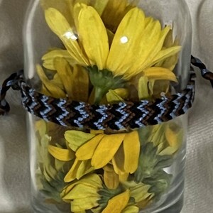 May include: A clear glass bottle filled with yellow flowers, a cork stopper, and a black cap. A woven bracelet with blue, brown, and white patterns encircles the bottle. The bottle is a decorative item.