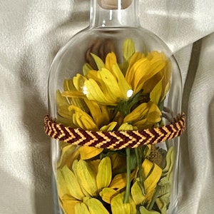 May include: A clear glass bottle filled with yellow flowers and petals. A braided bracelet in shades of brown and gold encircles the bottle. The bottle has a black cap and a cork stopper.