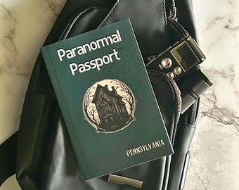 May include: A dark green "Paranormal Passport" book with a haunted house illustration and the word "Pennsylvania" is displayed on a black bag. A black digital recorder is partially visible in the bag's pocket.