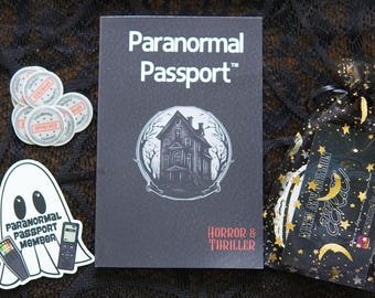 Puede incluir: Un libro negro "Paranormal Passport" con una ilustración de casa embrujada, texto "Horror & Thriller" y pegatinas. Una pegatina de fantasma con el texto "Paranormal Passport Member" y una bolsa de regalo negra con estrellas doradas.