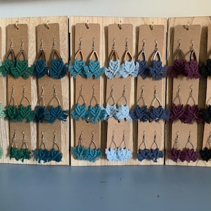May include: A display of handmade macrame earrings in various colors, including green, blue, and purple. Each pair features a unique design with a teardrop-shaped metal frame and a knotted fringe. The earrings are presented on individual cardboard displays.