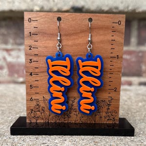 Op de afbeelding: Een paar blauwe en oranje acryl oorbellen met het woord "Illini" geschreven in een gestileerd lettertype.