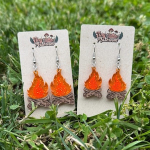 Könnte beinhalten: Zwei Paar Ohrhänger mit einem Lagerfeuer-Design. Jeder Ohrring hat eine orangefarbene Glitzerflamme über einer braunen Holzscheitbasis. Die Ohrringe werden auf einer beigefarbenen Karte mit dem Text "Tiny Mushroom" präsentiert.