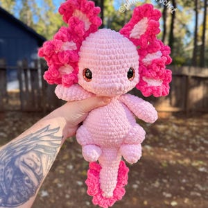 Puede incluir: Un juguete de peluche axolotl de crochet rosa con detalles blancos en las branquias. El juguete tiene ojos grandes y redondos y una cola larga y rizada. El juguete est&aacute; sostenido en una mano con un tatuaje.