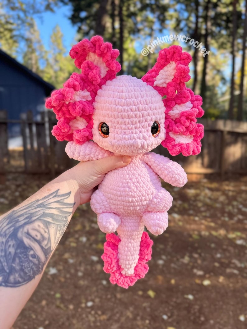 Puede incluir: Un juguete de peluche axolotl de crochet rosa con detalles blancos en las branquias. El juguete tiene ojos grandes y redondos y una cola larga y rizada. El juguete est&aacute; sostenido en una mano con un tatuaje.