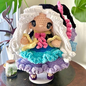 May include: A handmade crochet doll with a whimsical design. The doll features a light skin tone, large black eyes, and a two-tone hairstyle of white and black. It wears a colorful dress with layers of blue, green, and purple, and has pink and yellow accents.