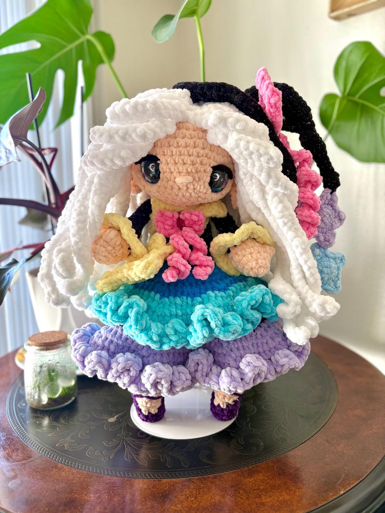 May include: A handmade crochet doll with a whimsical design. The doll features a light skin tone, large black eyes, and a two-tone hairstyle of white and black. It wears a colorful dress with layers of blue, green, and purple, and has pink and yellow accents.