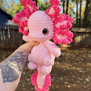 Puede incluir: Axolotl de peluche de crochet rosa con volantes rosa y blanco. El axolotl tiene ojos grandes y redondos con pupilas negras y un cuerpo rosa.