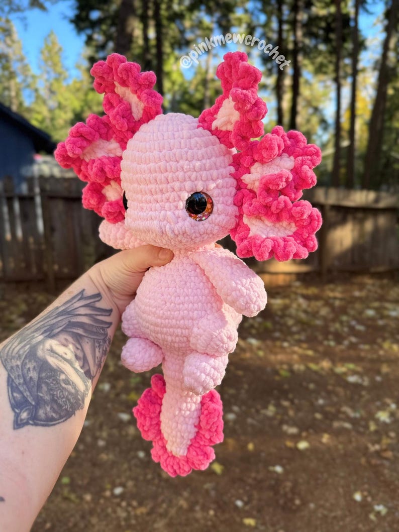 Puede incluir: Axolotl de peluche de crochet rosa con volantes rosa y blanco. El axolotl tiene ojos grandes y redondos con pupilas negras y un cuerpo rosa.