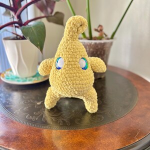 May include: A handmade, yellow crochet creature with iridescent eyes. The plush toy has a rounded body, small arms and legs, and a curved head. It is sitting on a dark brown, ornate table, with plants in the background.