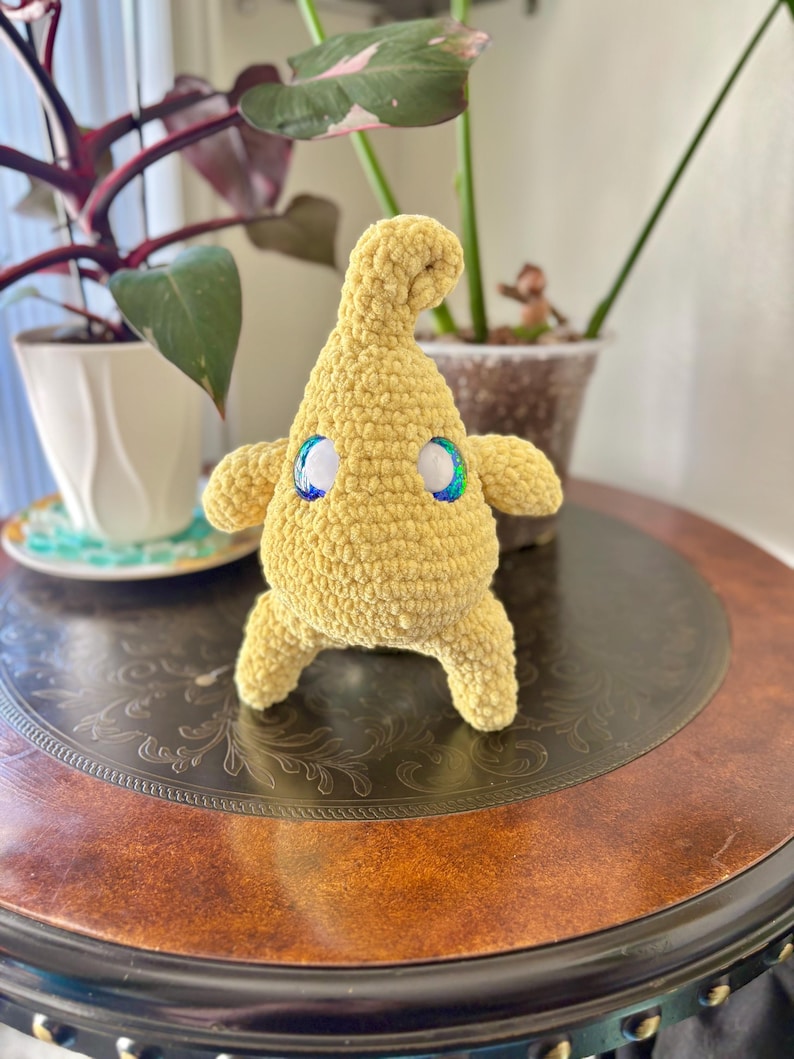May include: A handmade, yellow crochet creature with iridescent eyes. The plush toy has a rounded body, small arms and legs, and a curved head. It is sitting on a dark brown, ornate table, with plants in the background.