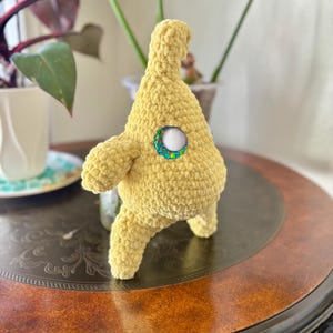 May include: A handmade, yellow crocheted creature with a button eye. The plush toy has a rounded body, small arms, and three legs. The button eye is white with a green and blue glittery border. The toy is sitting on a dark brown table.