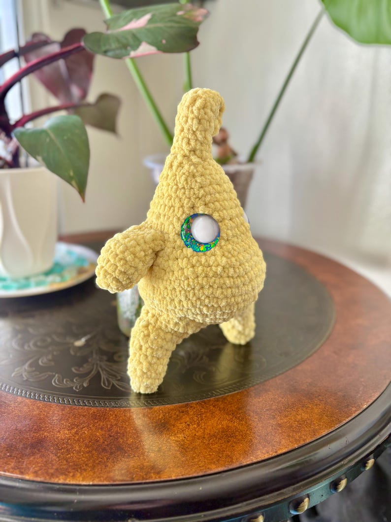 May include: A handmade, yellow crocheted creature with a button eye. The plush toy has a rounded body, small arms, and three legs. The button eye is white with a green and blue glittery border. The toy is sitting on a dark brown table.