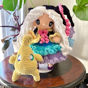 May include: A handmade crochet doll with white and black hair, wearing a colorful dress with blue, purple, and pink accents. A smaller yellow crochet creature with blue and green eyes stands beside the doll. Both are on a dark wooden table.