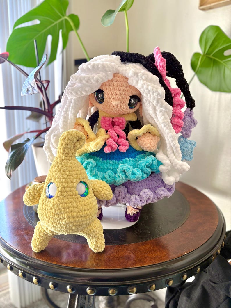 May include: A handmade crochet doll with white and black hair, wearing a colorful dress with blue, purple, and pink accents. A smaller yellow crochet creature with blue and green eyes stands beside the doll. Both are on a dark wooden table.