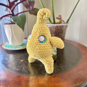 May include: A handmade, yellow crochet creature with large, round eyes featuring a white center and a colorful outer ring. The plush toy has small arms and legs, and a rounded head. It is displayed on a dark brown table with a decorative pattern.