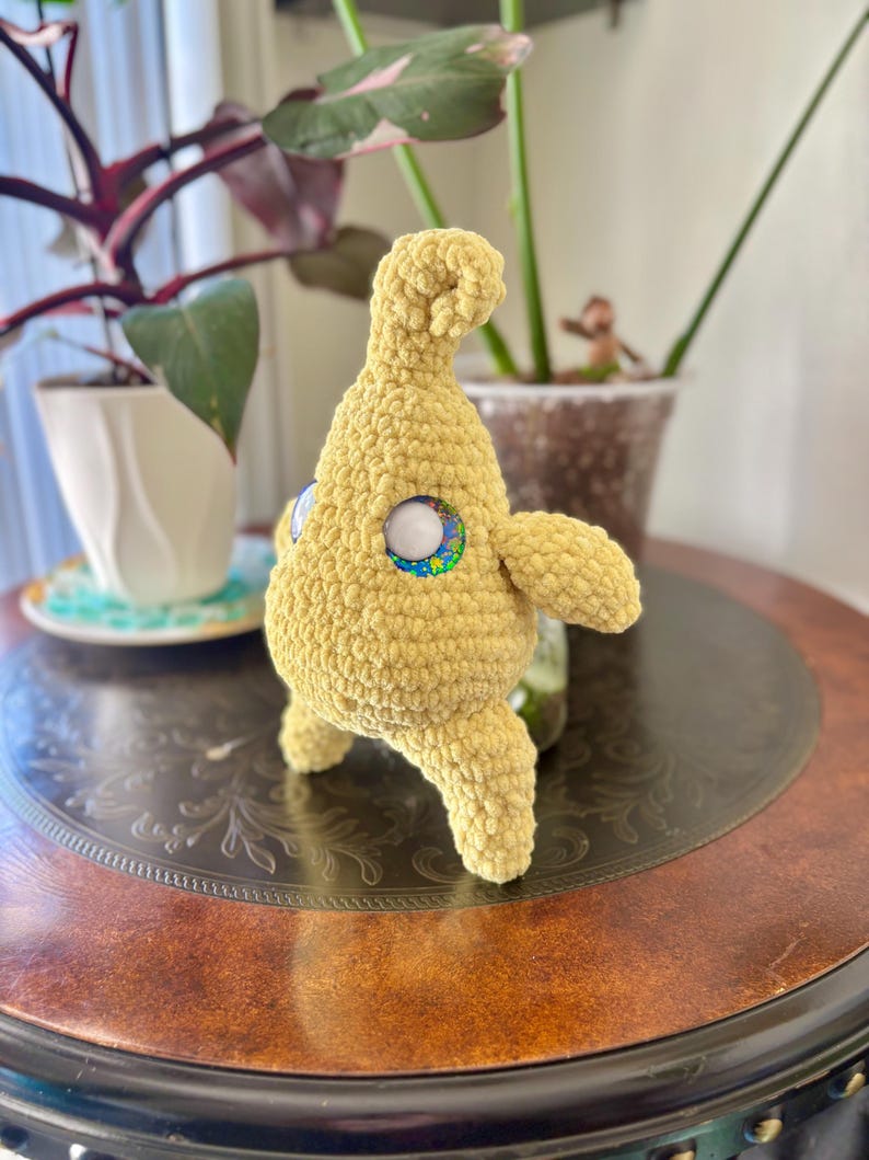 May include: A handmade, yellow crochet creature with large, round eyes featuring a white center and a colorful outer ring. The plush toy has small arms and legs, and a rounded head. It is displayed on a dark brown table with a decorative pattern.