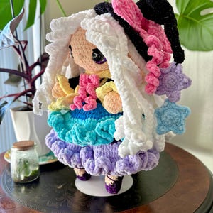 May include: A handmade crochet doll with white, fluffy hair and a colorful dress. The doll features a pink and black bow, a star-shaped accessory, and a multi-colored skirt in shades of blue, green, and purple. The doll is standing on a small white platform.