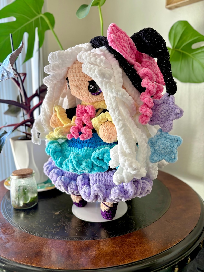 May include: A handmade crochet doll with white, fluffy hair and a colorful dress. The doll features a pink and black bow, a star-shaped accessory, and a multi-colored skirt in shades of blue, green, and purple. The doll is standing on a small white platform.