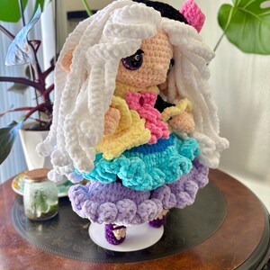 May include: A handmade crochet doll with a white, fluffy wig and a black hat. The doll wears a layered dress in yellow, pink, blue, and purple. The doll is standing on a white base with purple shoes. The doll is a unique, handcrafted item.
