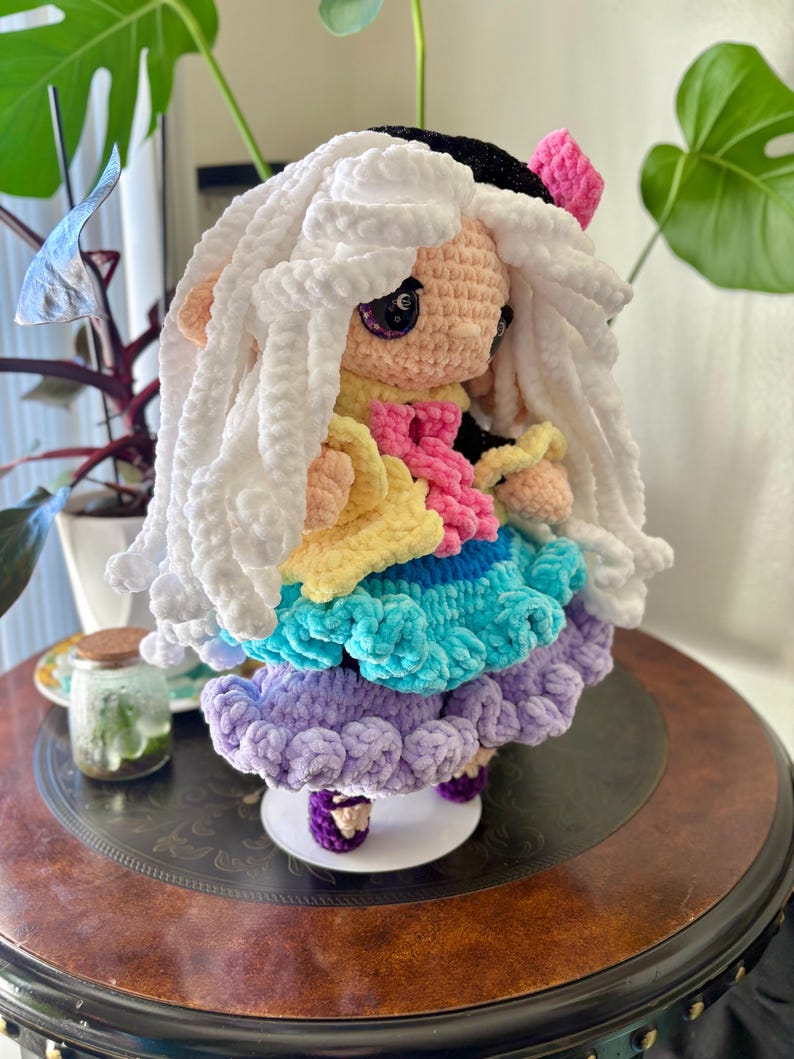 May include: A handmade crochet doll with a white, fluffy wig and a black hat. The doll wears a layered dress in yellow, pink, blue, and purple. The doll is standing on a white base with purple shoes. The doll is a unique, handcrafted item.