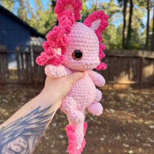 Puede incluir: Un juguete de peluche axolotl de crochet rosa con una cola rosa con volantes y volantes rosas alrededor de la cabeza. El axolotl tiene grandes ojos marrones y est&aacute; siendo sostenido por una mano con un tatuaje.