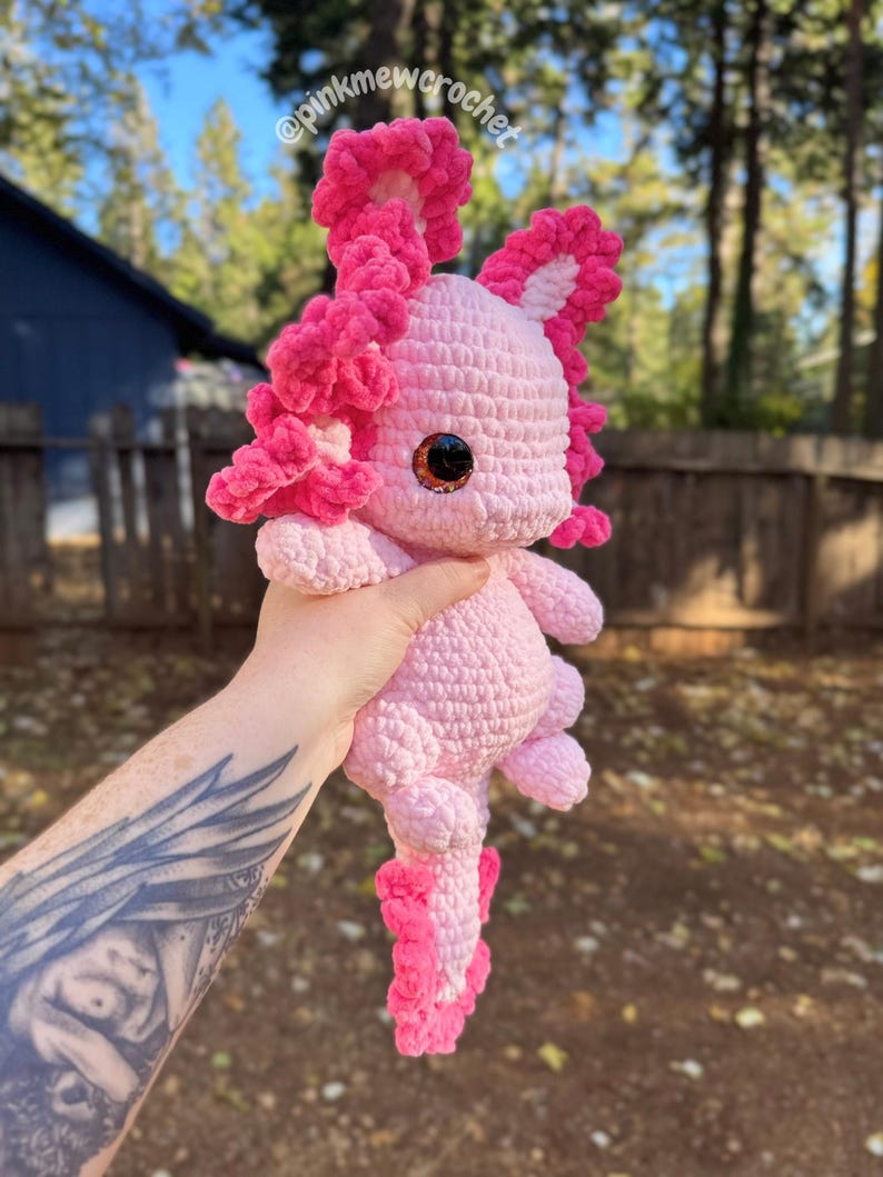 Puede incluir: Un juguete de peluche axolotl de crochet rosa con una cola rosa con volantes y volantes rosas alrededor de la cabeza. El axolotl tiene grandes ojos marrones y est&aacute; siendo sostenido por una mano con un tatuaje.