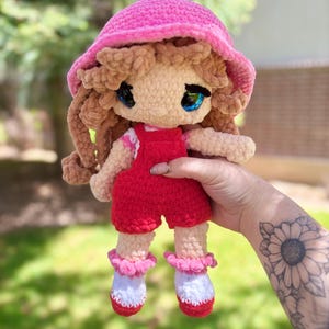 May include: A handmade crochet doll with a pink hat and red overalls. The doll has brown curly hair, blue eyes, and white and red shoes with pink ruffles. The doll is held by a person with a sunflower tattoo.