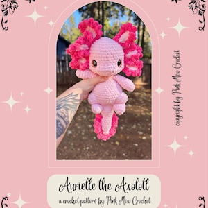 Puede incluir: Un juguete de peluche axolotl de crochet rosa con volantes rosas alrededor de la cabeza y la cola. El juguete est&aacute; siendo sostenido por una mano con un tatuaje. El texto "Aurielle the Axolotl" y "a crochet pattern by Pink Mew Crochet" est&aacute; en la parte inferior de la imagen.