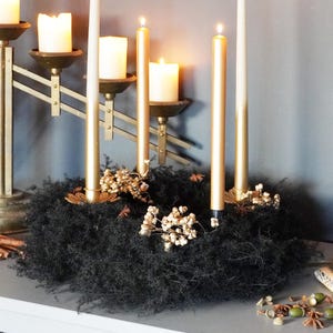Può includere: Una composizione festosa con candele accese di varie altezze e stili. Il centrotavola include una ghirlanda di muschio nero adornata con piccoli fiori bianchi e dettagli dorati. Sono presenti candelabri dorati e candele aggiuntive.