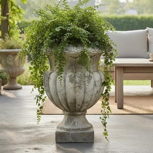 Könnte beinhalten: Eine große, verwitterte Steinvase, die mit grünen Pflanzen überquillt. Die Vase hat ein detailliertes, klassisches Design und steht auf einem quadratischen Sockel. Der Hintergrund umfasst eine Holzpergola, Sitzgelegenheiten im Freien und anderes Grün, wodurch eine ruhige Gartenszene entsteht.