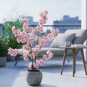 Op de afbeelding: Een kunstmatige kersenbloesemboom in een pot met lichtroze bloemen. De boom staat in een grijze, getextureerde pot en staat op een terras. Een ligstoel, een bijzettafel en een stadsgezicht zijn op de achtergrond.