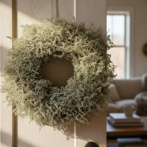Op de afbeelding: Een ronde krans van gedroogd mos hangt aan een lichtgekleurde houten deur. De krans is lichtgroen van kleur en heeft een dichte, getextureerde uitstraling. De achtergrond toont een woonkamer met een bank en salontafel.