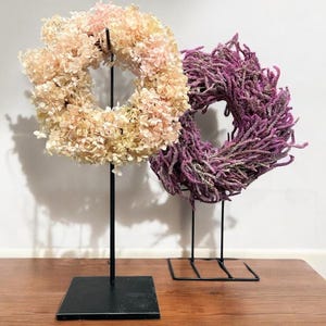 Könnte beinhalten: Zwei dekorative Kränze auf schwarzen Metallständern. Ein Kranz besteht aus hellrosa und cremefarbenen Blumen. Der andere Kranz besteht aus violettem, strukturiertem Material. Beide Kränze sind kreisförmig und werden auf einer Holzoberfläche ausgestellt.