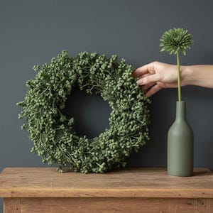 Può includere: Una ghirlanda circolare composta da numerosi piccoli grappoli verdi testurizzati simili a bacche. Accanto alla ghirlanda si trova un vaso opaco a forma di bottiglia color verde oliva contenente un fiore verde con una corolla spinosa.