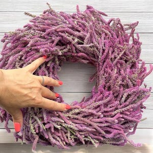 Könnte beinhalten: Ein quadratischer Kranz aus getrockneten, strukturierten, lila Blumen. Eine Hand mit rotem Nagellack und einem silbernen Ring berührt den Kranz. Der Hintergrund ist eine hellgraue Holzoberfläche.
