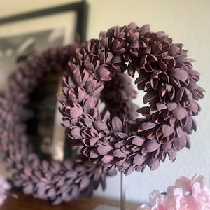 Könnte beinhalten: Zwei dekorative, kreisförmige Kränze aus kleinen, dunkelvioletten, blütenblattartigen Elementen. Die Kränze haben eine strukturierte Oberfläche. Der Hintergrund zeigt ein gerahmtes Schwarz-Weiß-Bild und rosa Blumen.