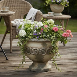 Puede incluir: Una gran jardinera beige ornamentada llena de una variedad de flores coloridas y hiedra colgante. La jardinera tiene un diseño de relieve detallado y se encuentra en una terraza de madera. Otras jardineras y muebles de mimbre están en el fondo.