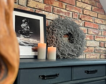 May include: A black framed photo of a rock stack on a snowy landscape, two white pillar candles, and a gray dried flower wreath on a dark gray dresser with two drawers.