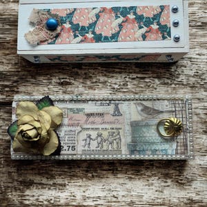May include: Two decorative wooden boxes. The top box is white with a patterned design of women in dresses, a blue bead, and lace. The bottom box has a paper collage design, a gold rose, and a pearl border.