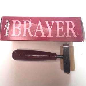 Puede incluir: Un rodillo Speedball BRAYER granate con un mango granate oscuro y un rodillo beige. La caja es granate con la palabra "BRAYER" en letras blancas grandes.