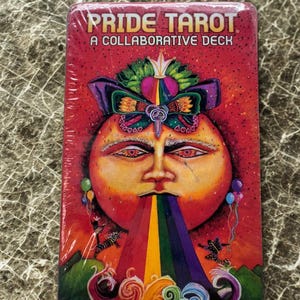 May include: A deck of Pride Tarot cards. The box is red and features the words "PRIDE TAROT" and "A COLLABORATIVE DECK". The artwork depicts a face with a rainbow coming from its mouth. There are also colourful balloons and swirls.