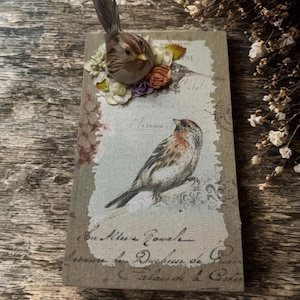 May include: A decorative journal with a vintage bird illustration and a 3D bird embellishment. The journal features a printed design with script and floral accents. The bird is brown with a long feather tail. The journal is approximately 6 inches tall.