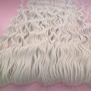 Puede incluir: Una pieza de extensión de cabello largo y ondulado en color blanco brillante. El cabello presenta un patrón de ondas profundas y consistentes en toda su longitud, con una base de malla fina y transparente visible en el borde superior de fijación.