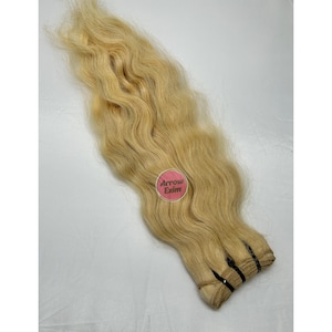 May include: Blonde wavy hair extensions. The hair is a light, golden color and appears to be human hair. The extensions are bundled together at the bottom with black bands. The image includes a pink sticker with the text "Arrow Exim".