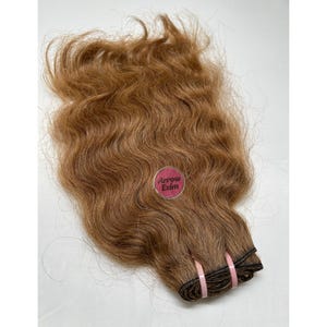 Op de afbeelding: Een bundel golvende, lichtbruine hairextensions. Het haar is aan de basis samengebonden met zwart garen en vastgezet met twee roze elastiekjes. Een ronde sticker met de tekst "Arrow Exim" is zichtbaar.