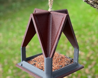 May include: A brown and gray bird feeder with a triangular roof and a square base. The feeder is hanging from a tree branch with a rope.